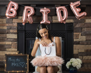 Young smiling woman opening a bridal shower present with pink bride balloons and brick fireplace in background