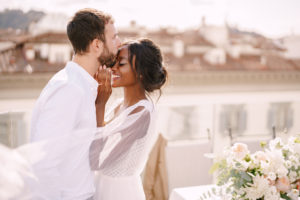 Destination fine-art wedding in Florence, Italy. Caucasian groom and African-American bride cuddling on a rooftop in sunset sunlight. Multiracial wedding couple